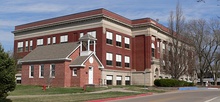 Small brick building with wood belltower in foreground; large three-story brick-and-stone building behind