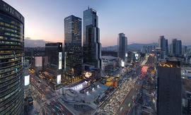 An evening rush hour shot of the intersection between the Teheran boulevard and Yeongdong highway from above. Multiple skyscrapers are visible, along with a pedestrian plaza and a stadium. In the background, apartment buildings and mountains are visible.