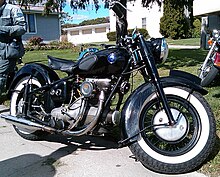 A mildly customized Sunbeam S7 motorcycle sits along the edge of a driveway. It shows evidence of normal wear and tear, but appears to be in very good condition. It is all black, with a vaguely eggplant-shaped fuel tank that features rubber pads for the rider's knees, and white-wall tires.