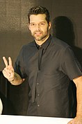 A man looking ahead, in a dark, half-sleeved shirt. He makes the 'V' sign gesture with his right hand.