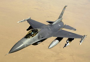 Aerial view of jet aircraft, carrying cylindrical fuel tanks and ordnance, overflying desert