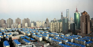 A cityscape seen from a height. In the foreground are low-rise buildings with blue roofs, giving way at the rear to higher-rising towers of different shapes and materials.