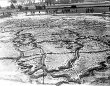 a vast full-scale model of the Vimy Ridge battlefield sprawls out across the landscape, resembling a football field in size. The intricate details of the terrain, including trenches, craters, and rough terrain, are meticulously recreated, providing a vivid representation of the historic battleground. Along the periphery of the model, soldiers stand, their figures adding a human dimension to the scene. The soldiers' presence shows the scale of the battlefield model.