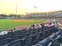 A view of a green baseball field from the third base side seats showing men in white baseball uniforms playing their positions with the late afternoon sun above the horizon