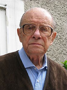 A bust photograph of an older white man; he has little hair, is wearing large glasses and looking into the camera unsmiling.