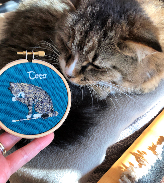 An embroidered likeness of a tabby cat on blue fabric in a small round frame, with the actual cat peering over the edge of the frame as if looking at the embroidery.