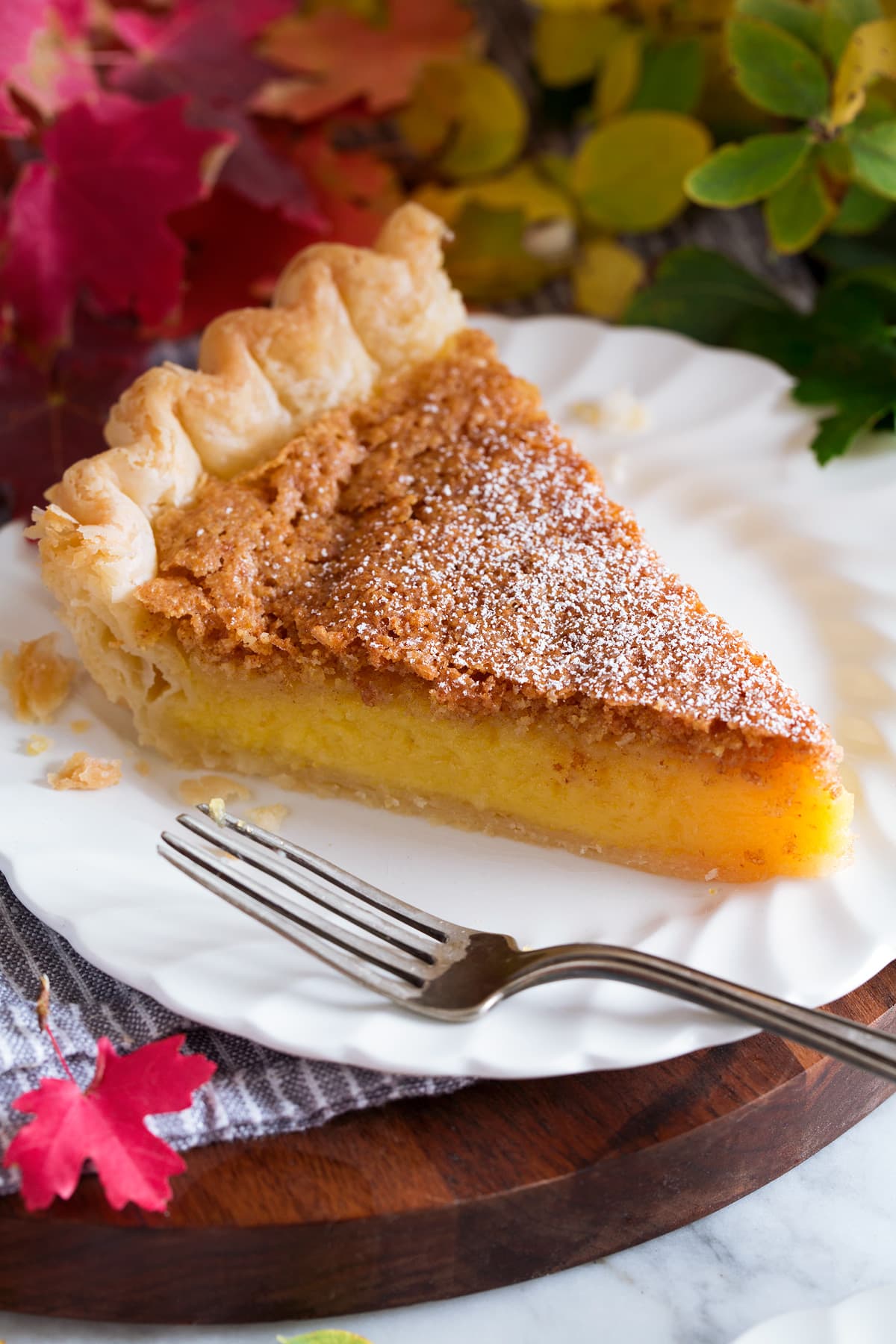 Slice of southern chess pie served on a white plate with scalloped edges. Fall foliage is shown surrounding the plate.