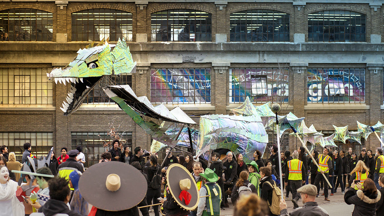 A metallic, green dragon with scales surrounded by students and staff.