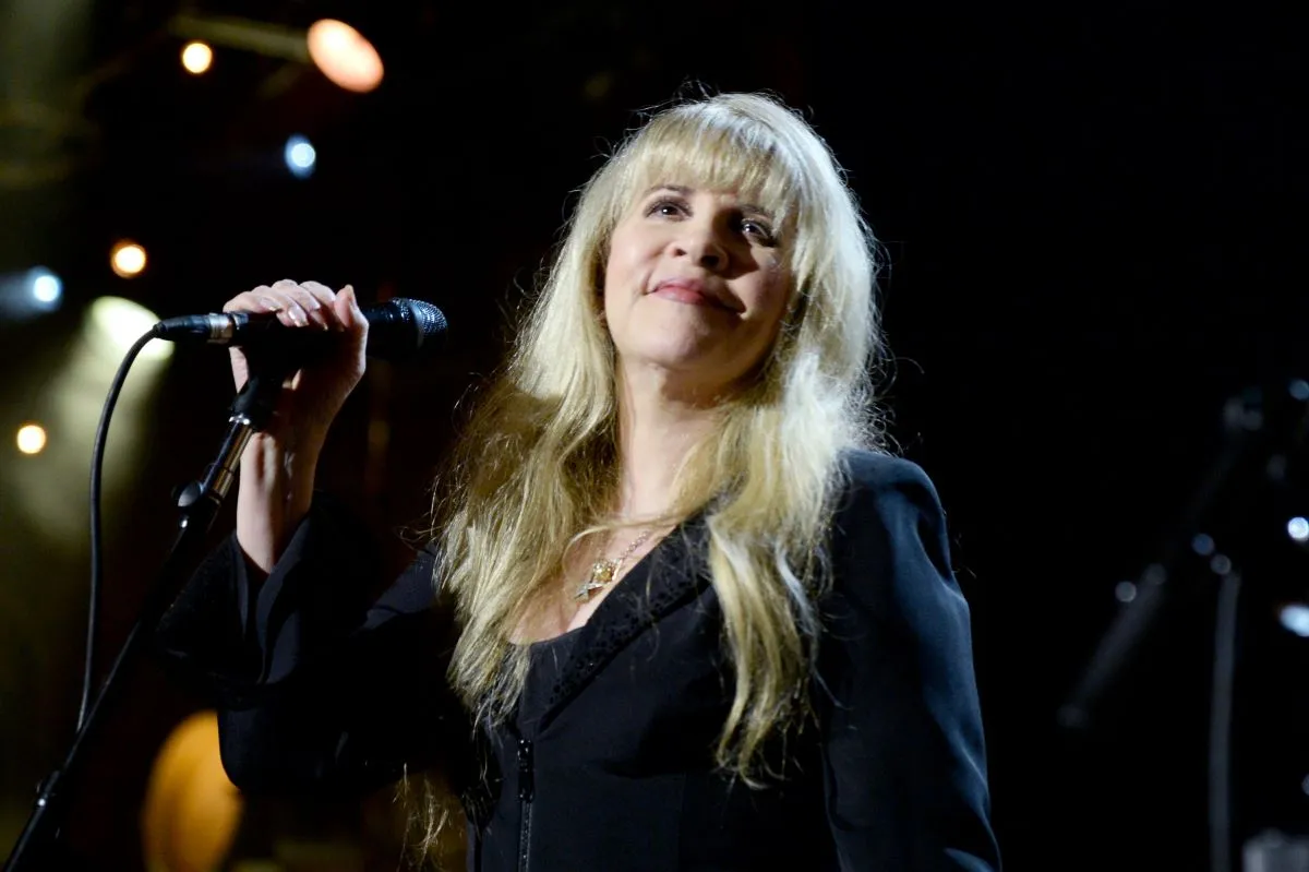 Stevie Nicks wears a black dress and holds a microphone in one hand.