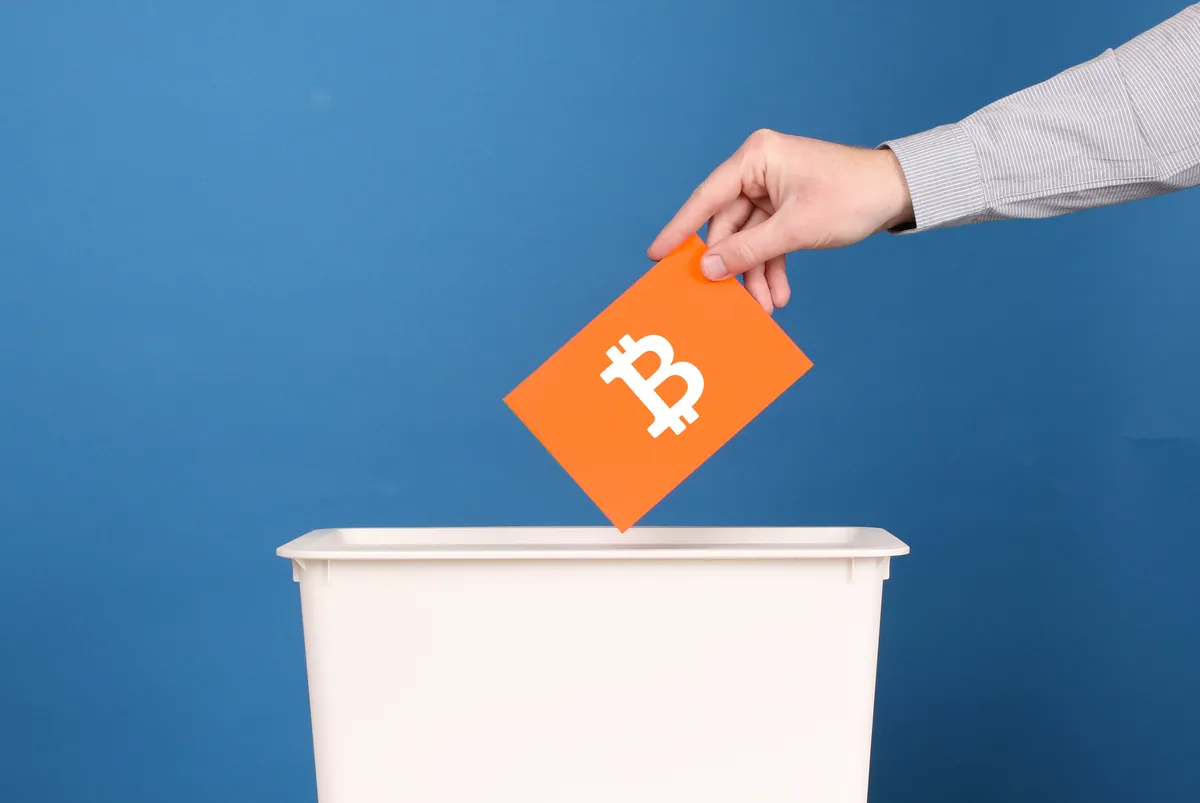 A person dropping an orange envelope with a white bitcoin symbol into a ballot box
