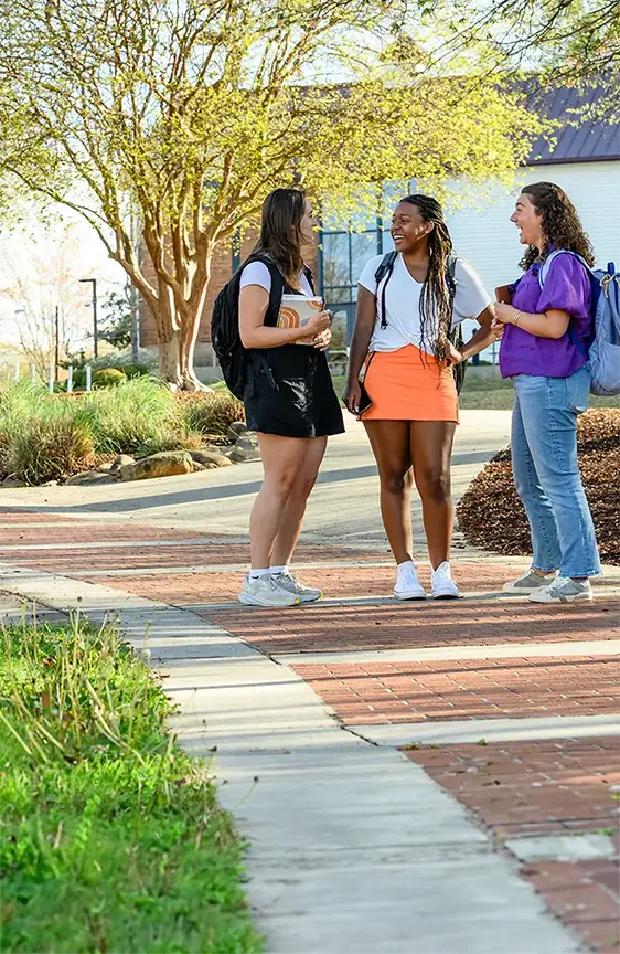 Three female students with backpacks stand on a walkway in the middle of campus in front of a painted white and brick building and green leafy trees.