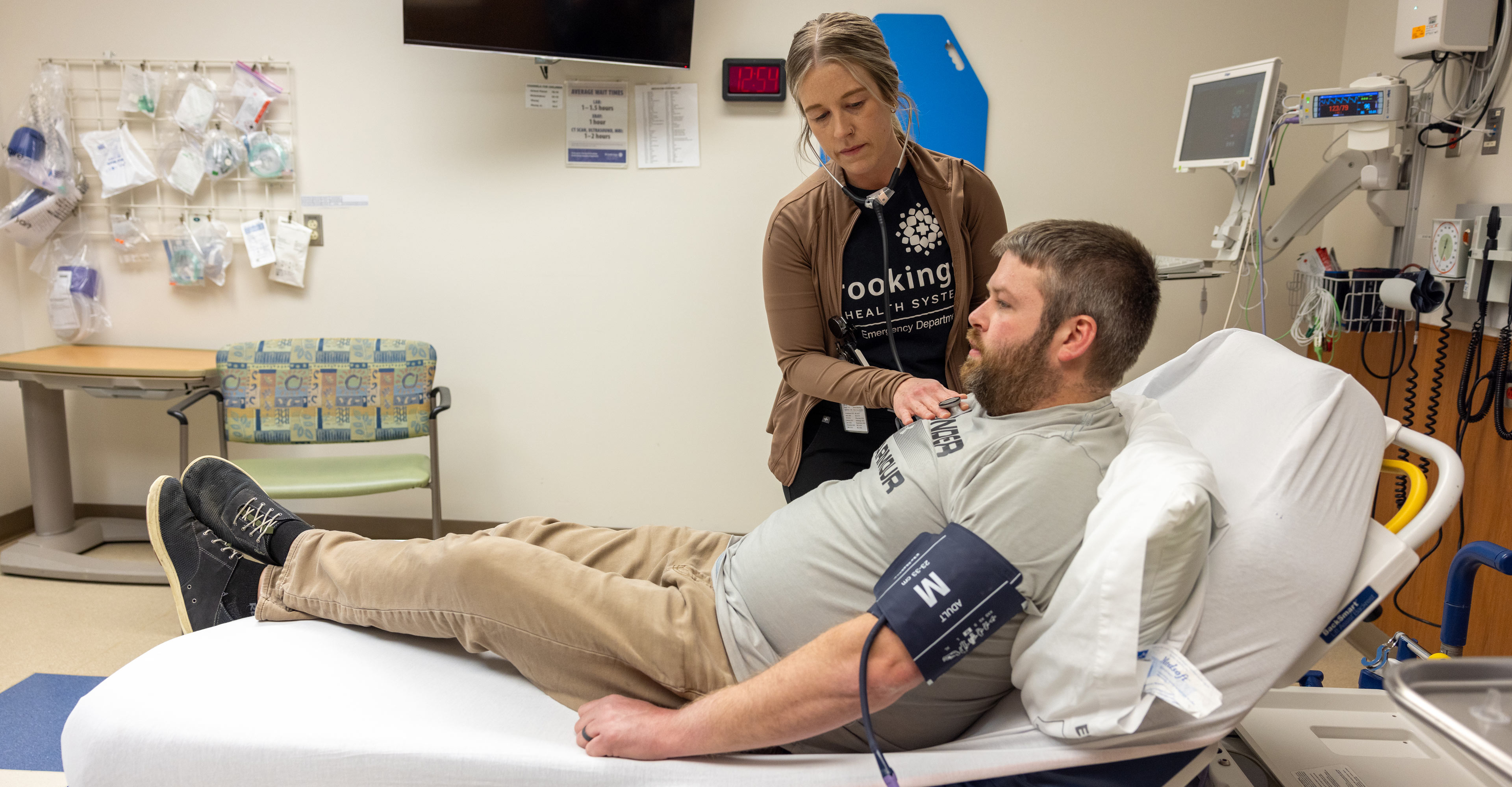 Female nurse using a stethoscope to listen to the lungs of a male patient who is reclined in a bed in an emergency patient room