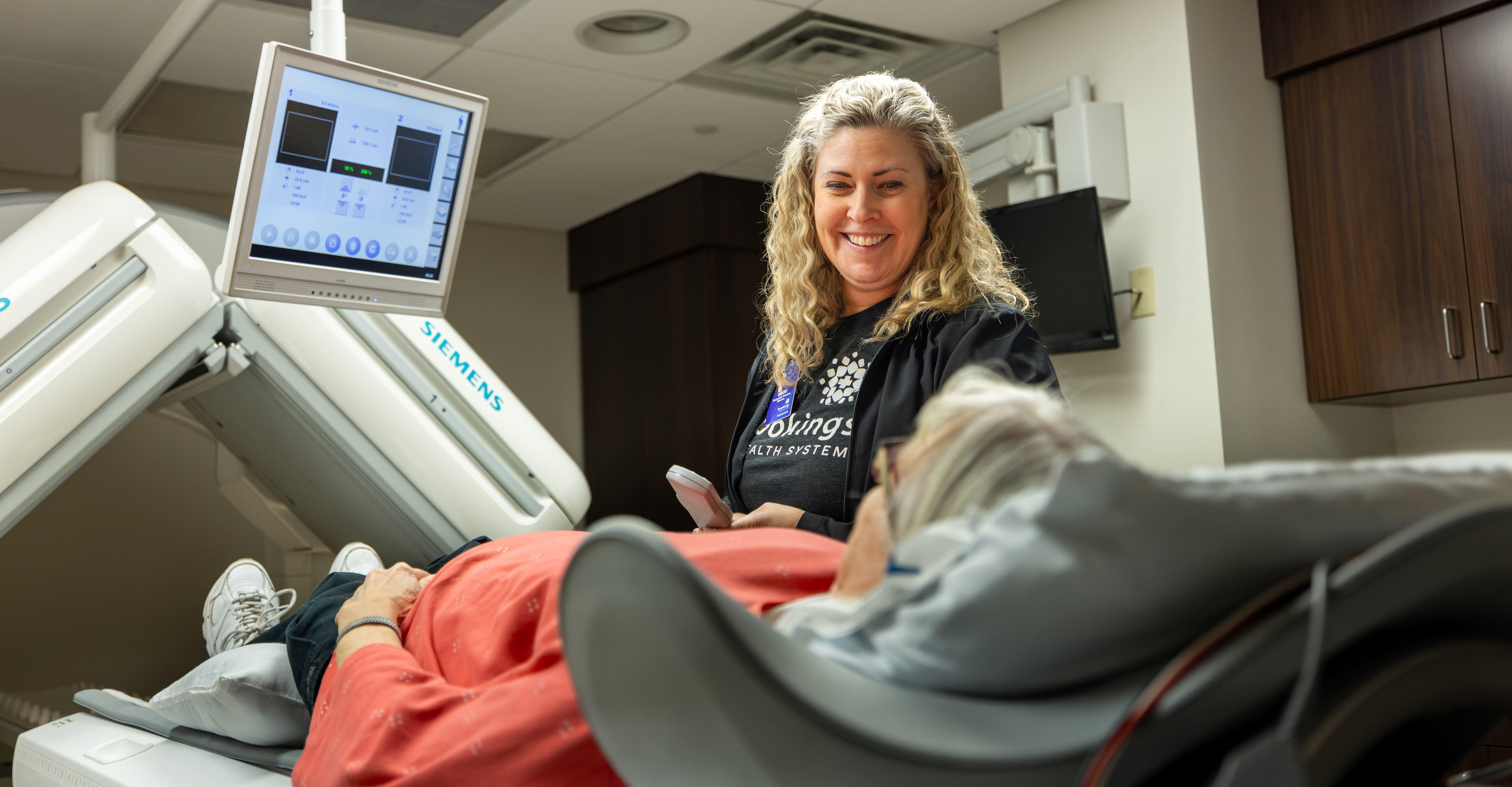 Female nuclear medicine technologist smiling at an older female patient lying on the bed, waiting to be scanned