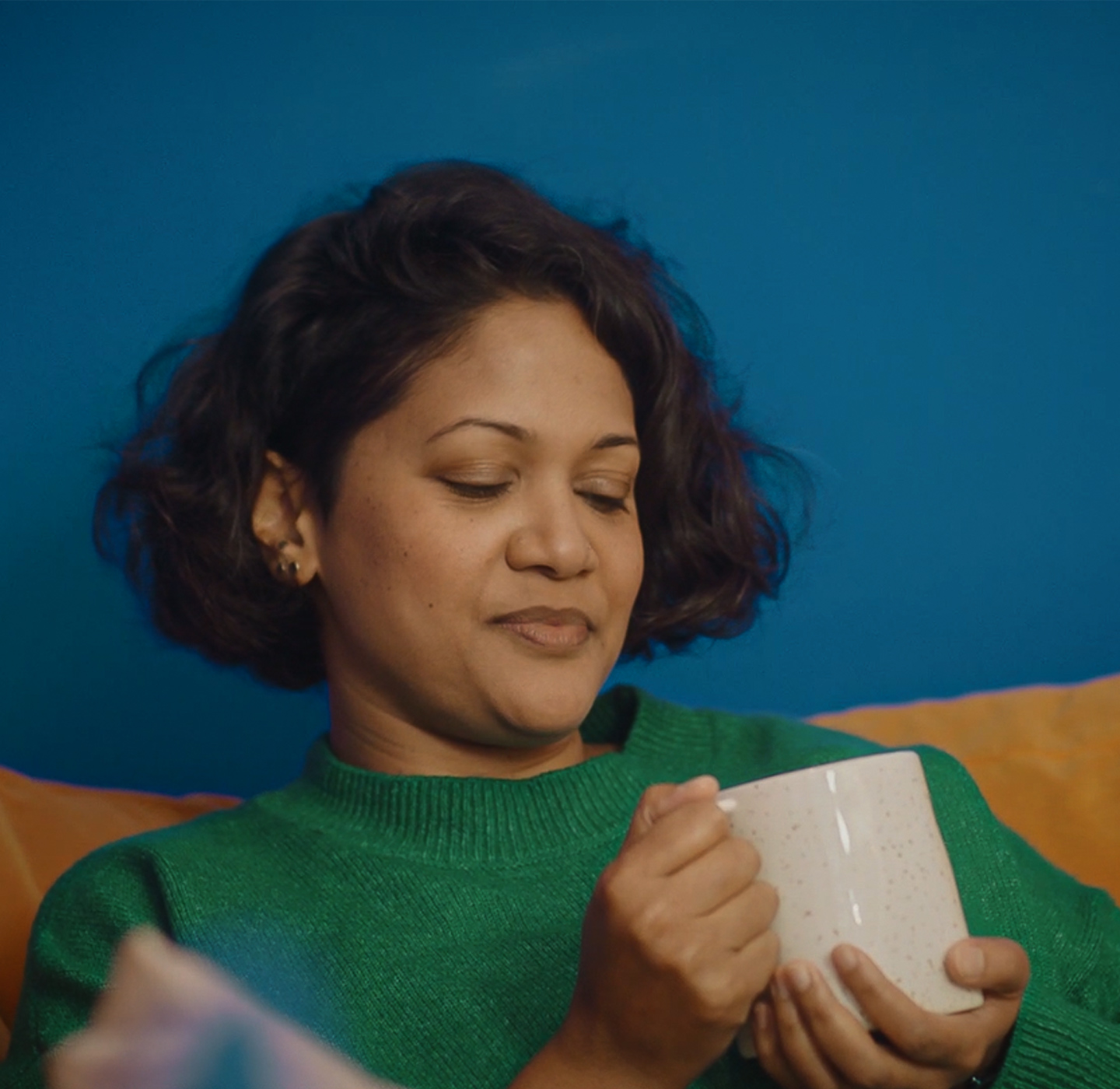 A young woman sat on a sofa looking at a cup of tea she is holding in her hands