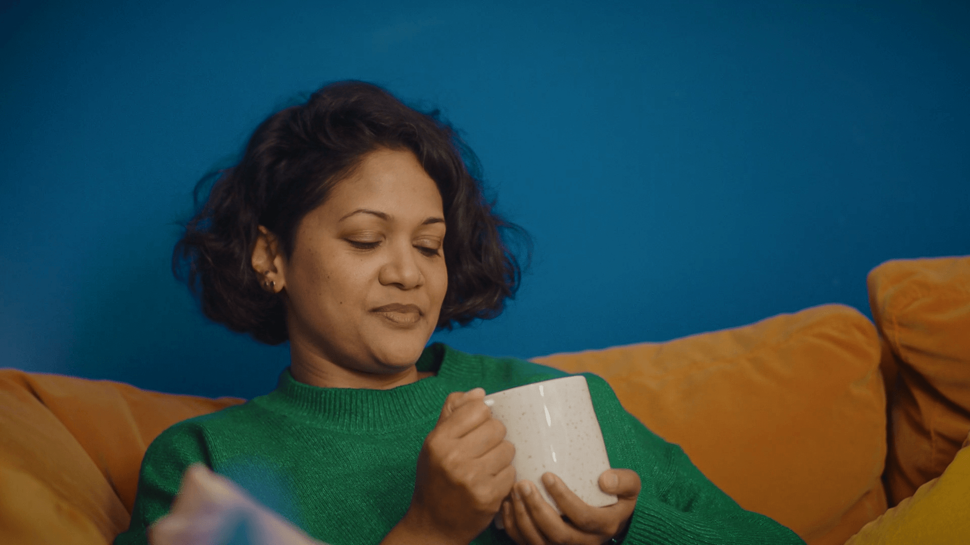 A young woman sat on a sofa looking at a cup of tea she is holding in her hands