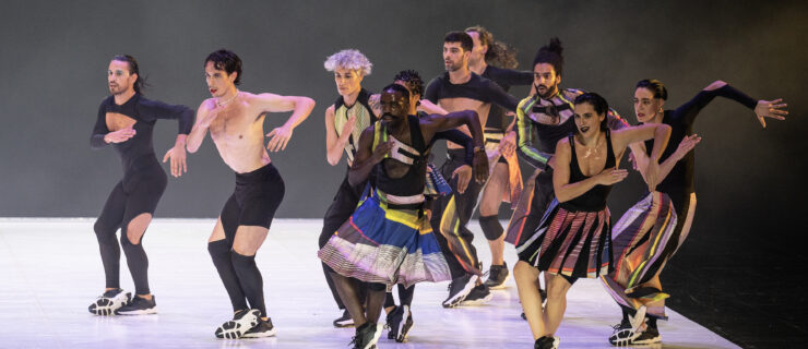 Ten dancers in various costumes of pieced together from athletic gear in black with brightly colored stripes move in unison. They cross one foot over the other, arms working in opposition as though they are running, or moving through a salsa step. All wear sneakers and are intently focused toward the downstage right corner.