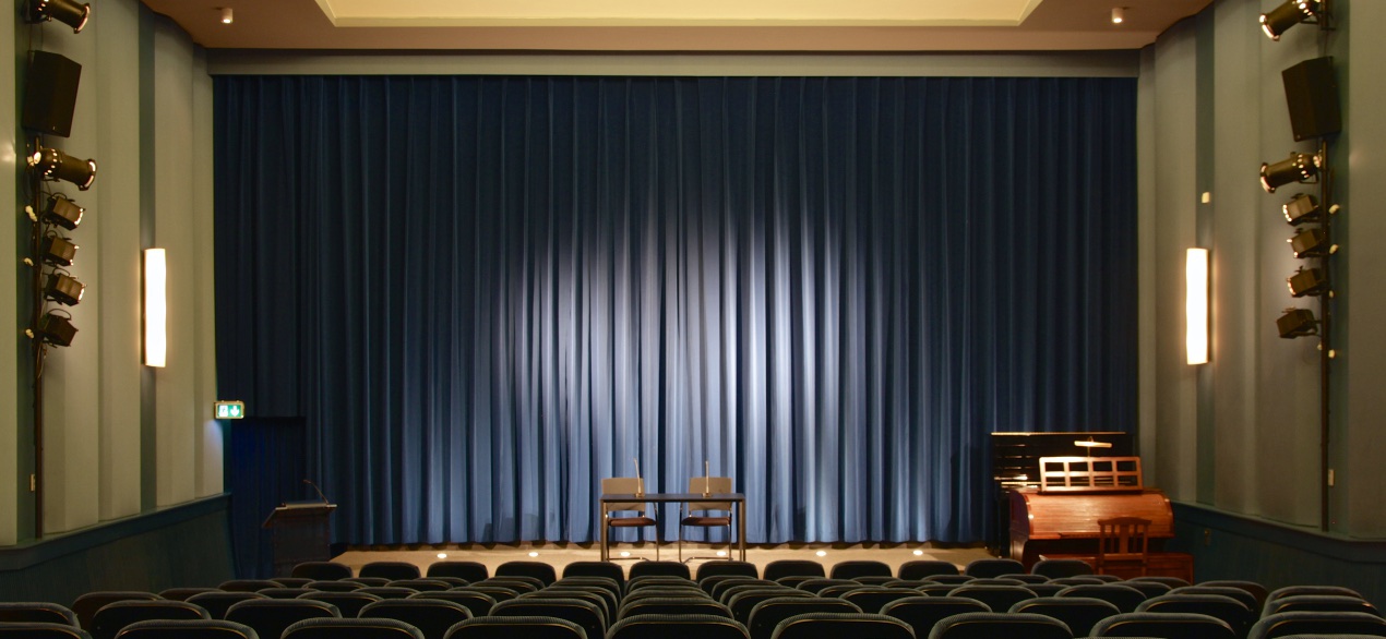 Das Bild zeigt den Kinosaal des Filmmuseum Potsdam. Im Detail ist ein blauer Vorhang der großen Leinwand sowie Bestuhlung und Beleuchtung zu sehen.