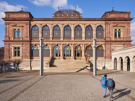 Vom Vorplatz aus wird die symetrische Vorderansicht des rot-bräunlich-backsteinfarbenen Museums mit seinem Treppenaufgang hin zum oberen Eingangsbereich gezeigt.