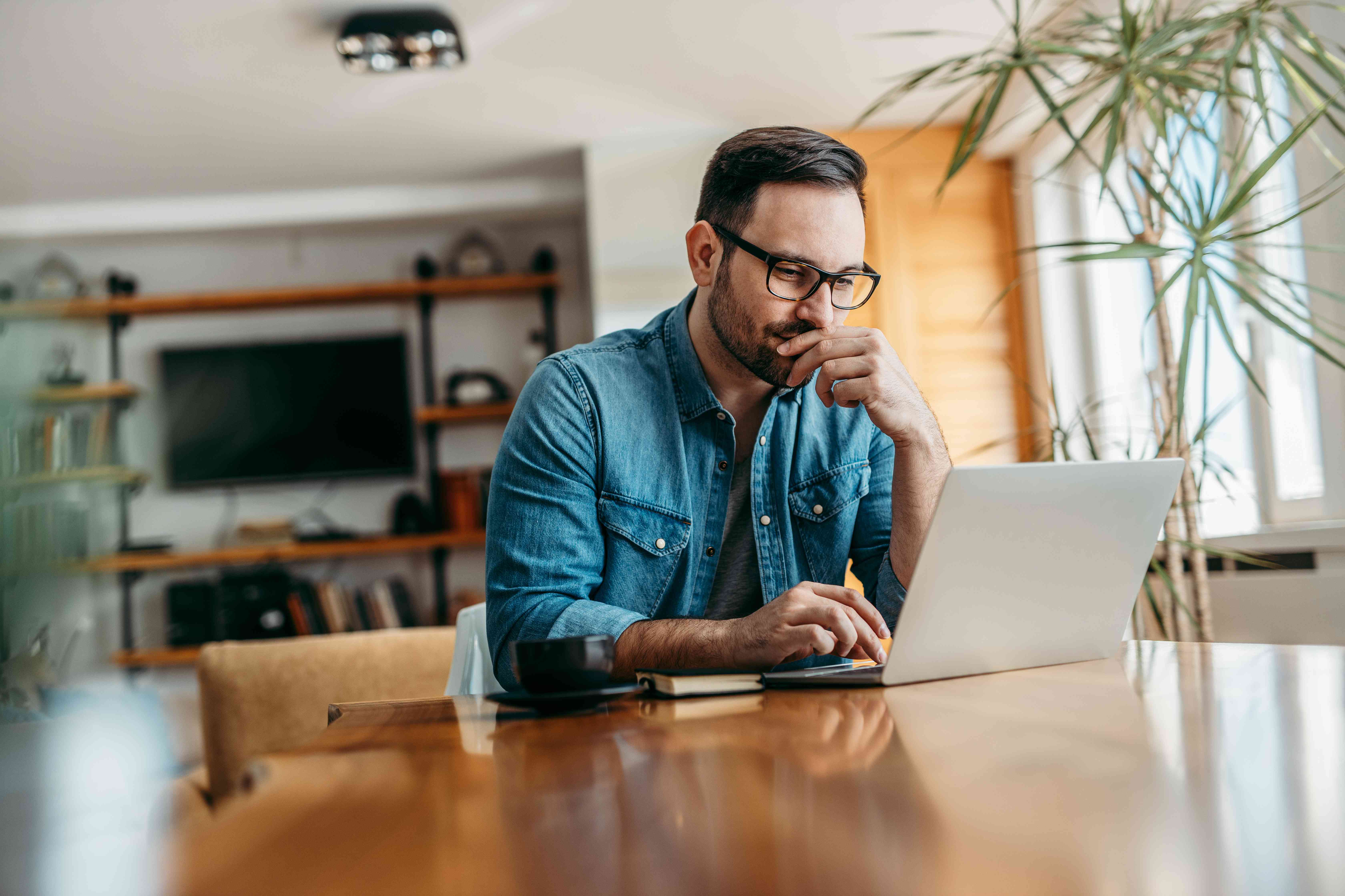 Man in his 30s at his dining room table looking carefully at his laptop screen