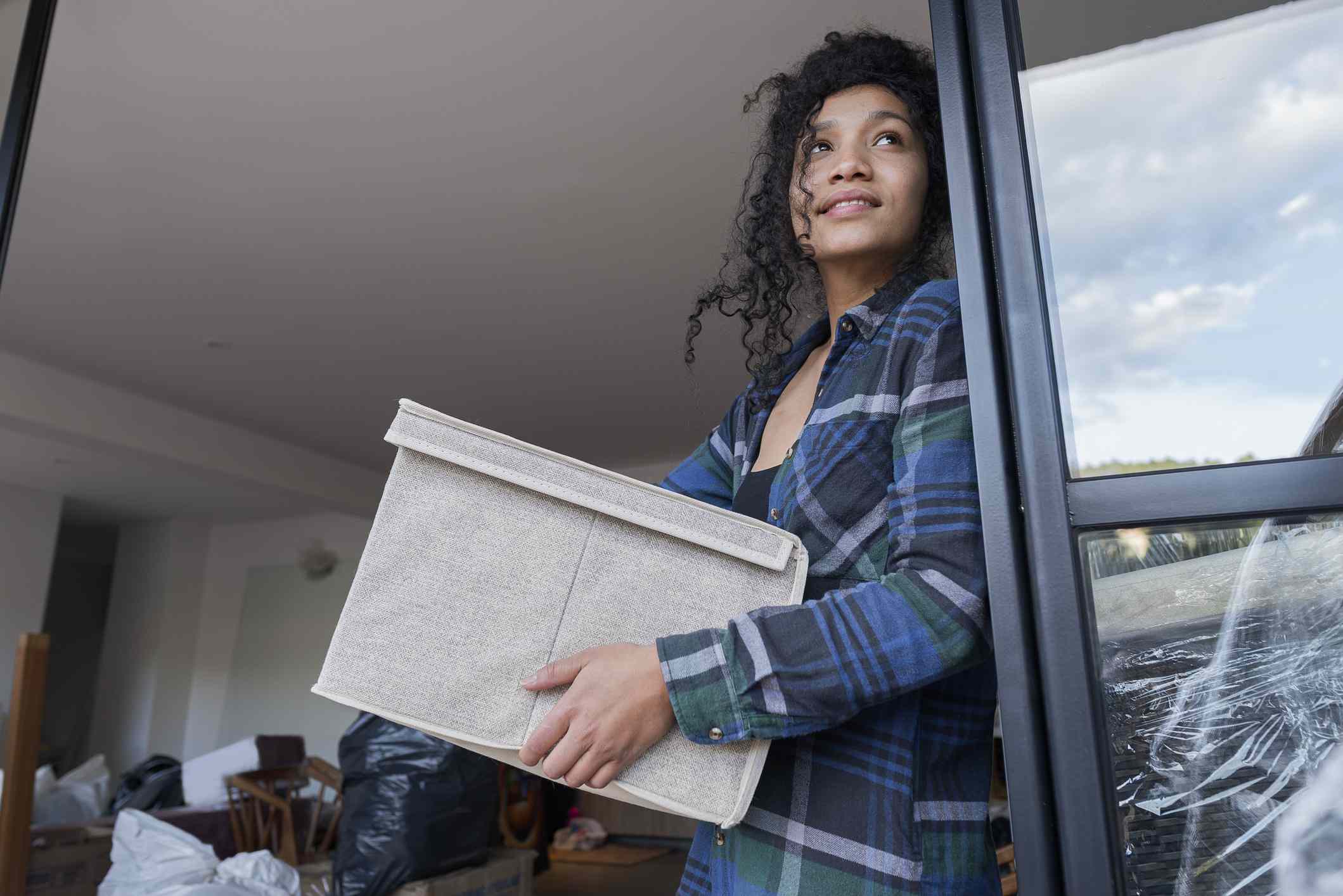 Young dark-haired adult holds a box while looking out the window of an apartment she is packing up ahead of moving. 