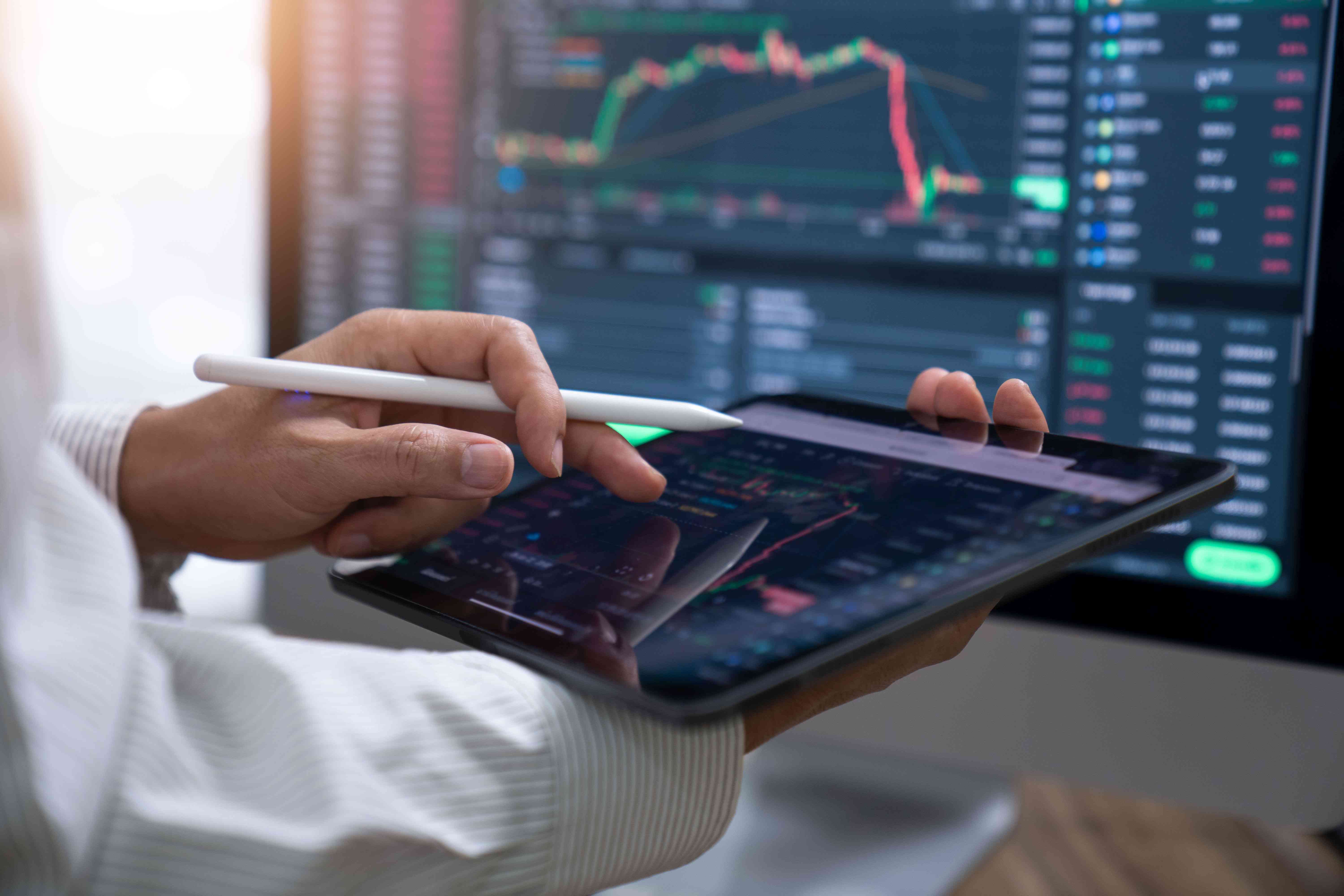 Investor using a tablet and computer screen to trade assets