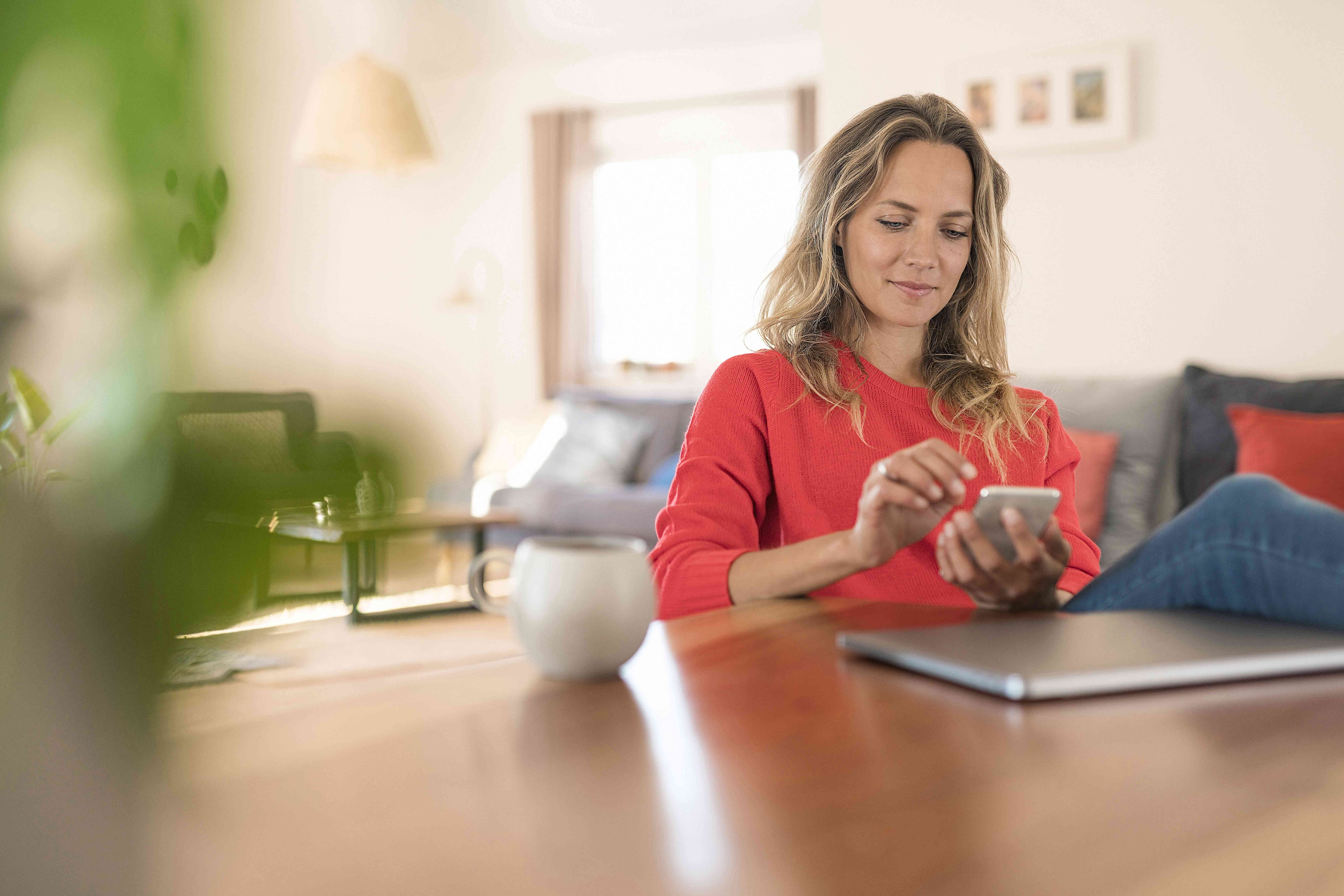 Woman in her 30s or 40s at her dining room table, looking at her smartphone with a smile, while a laptop sits nearby