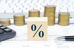 A wooden cube showing a percentage sign in front of varying height stacks of coins with a calculator, pen, and paperwork