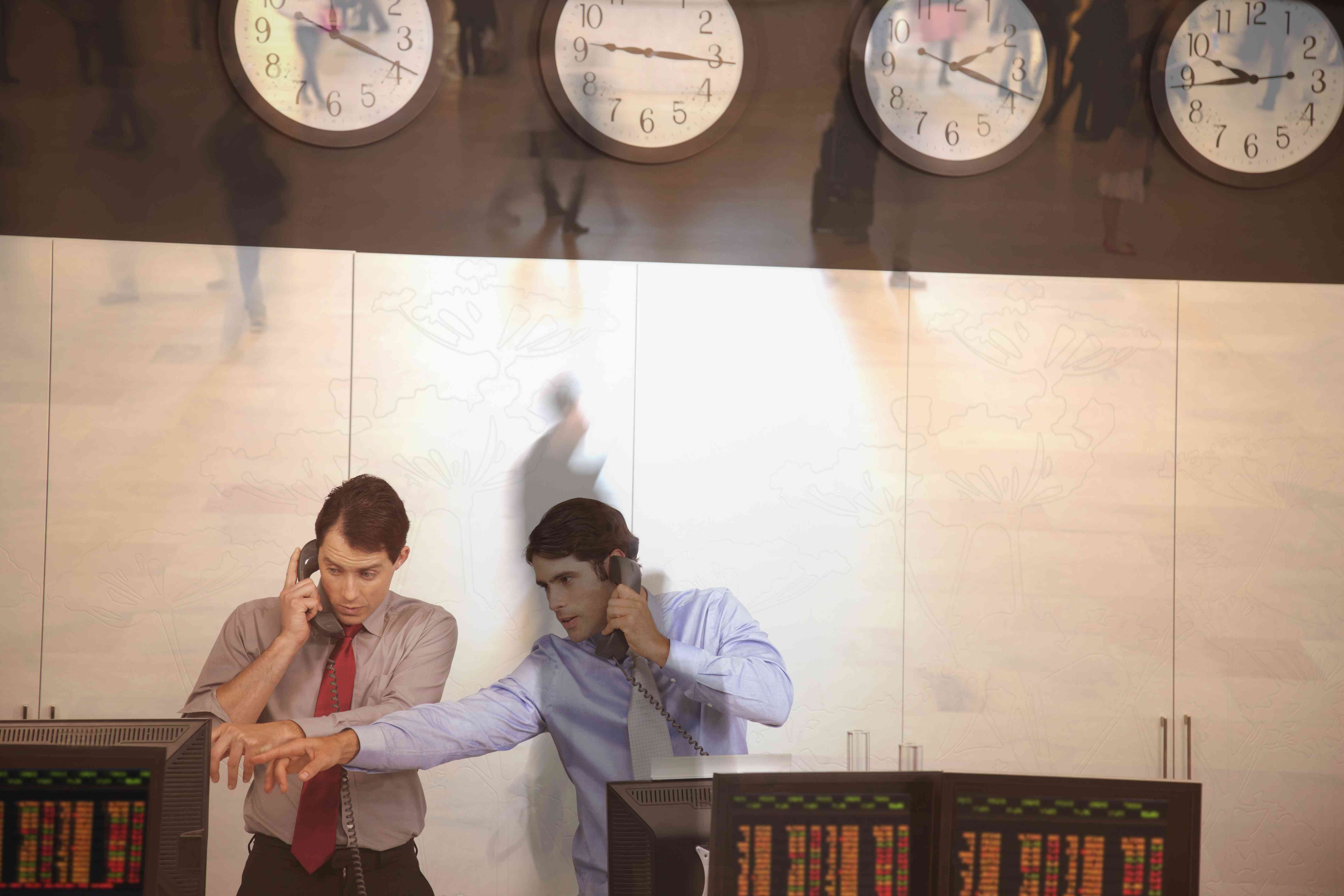 Two derivative traders stand behind trading consoles and take trade orders on the phone while above them a series of clocks display the time in different time zones.