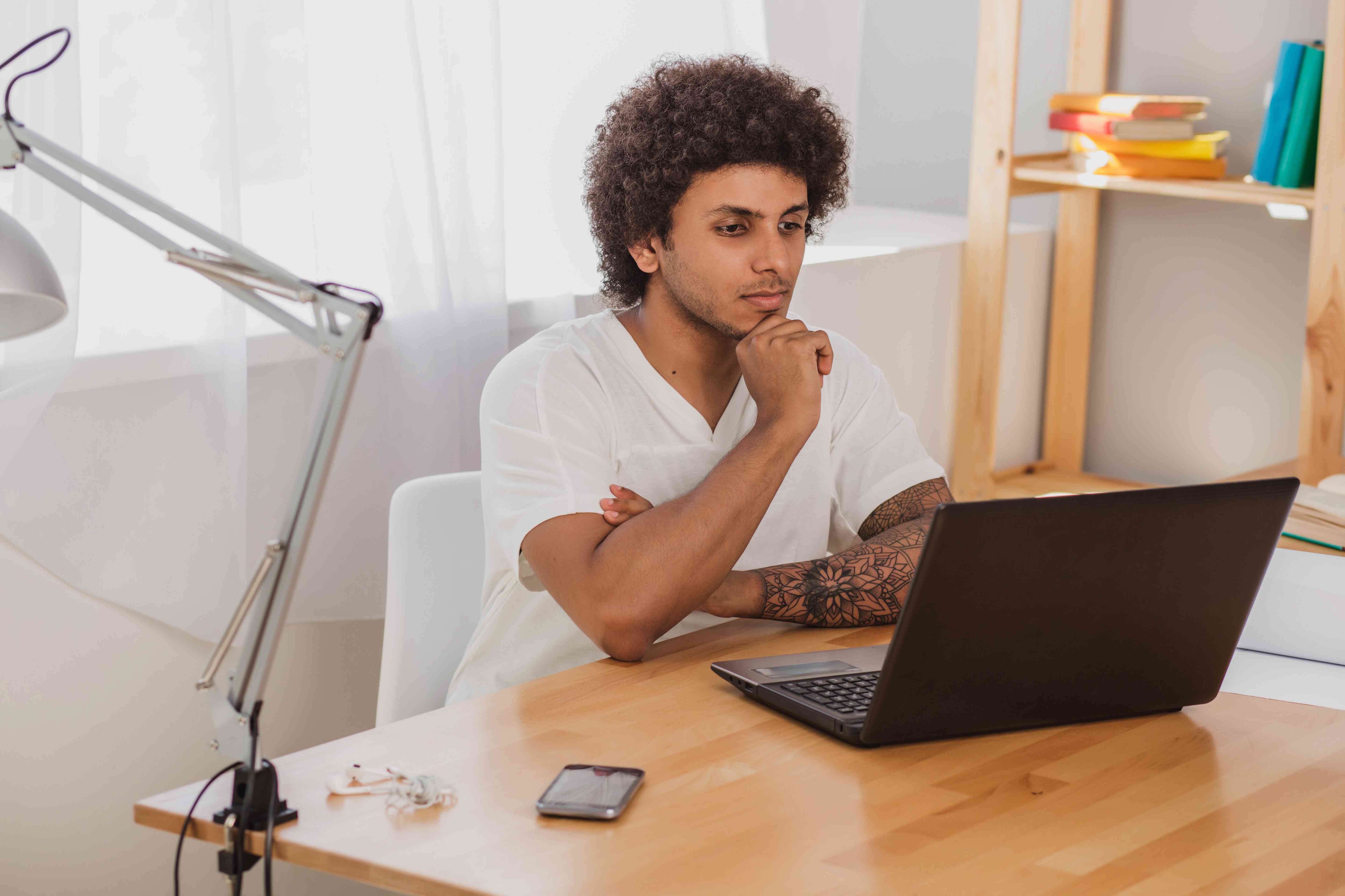 Young man sitting at a home desk and intently studying something on his laptop screen