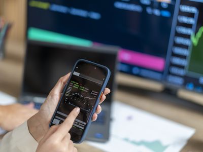 A woman holds a cell phone to check stock market prices, with a computer screen in the background.