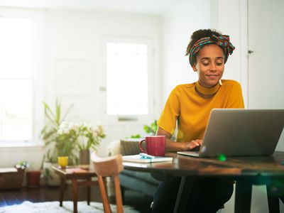 A woman in a yellow shirt sits at a table at home with a laptop and a red mug