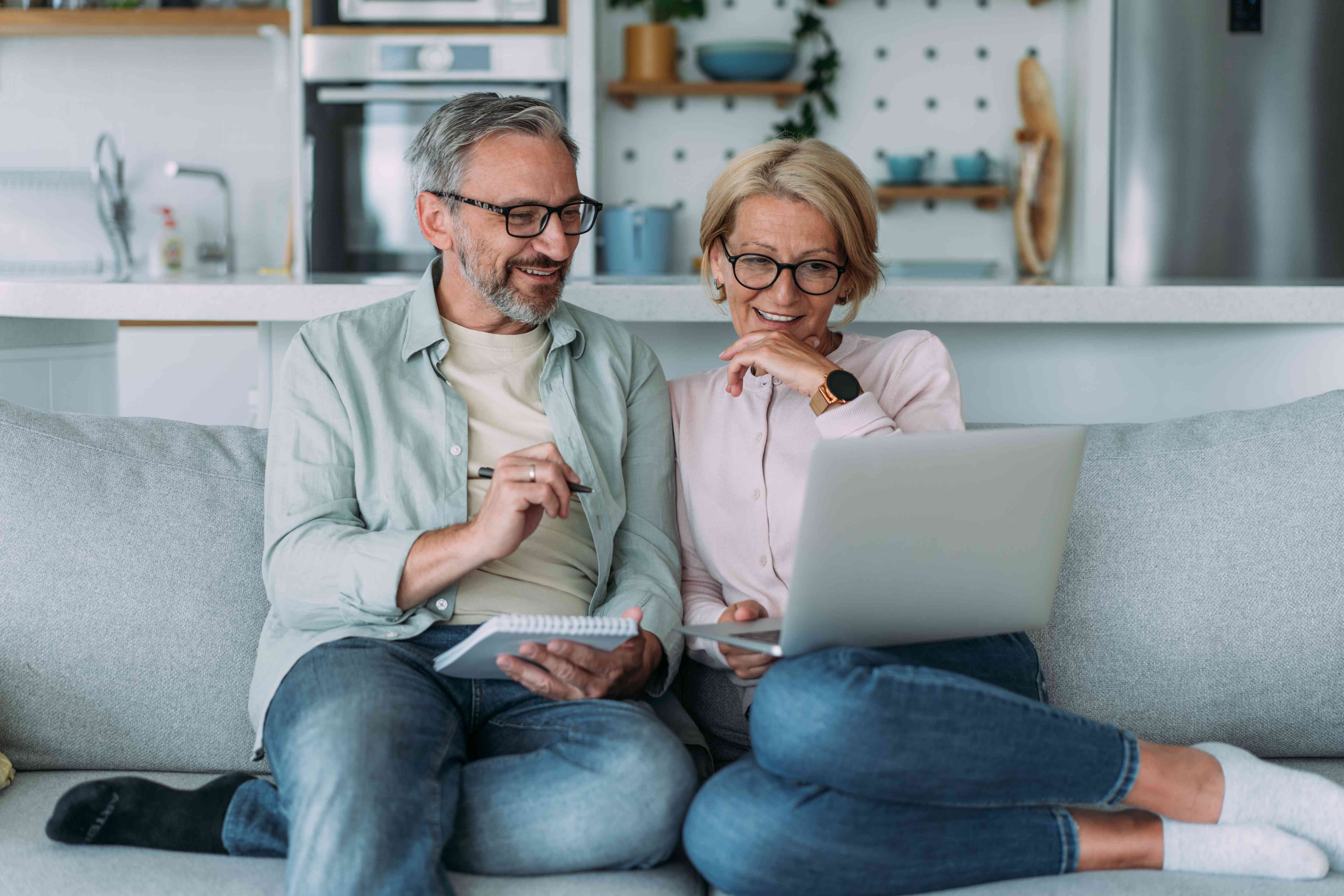 Older couple sitting together on their couch, with pen and paper and looking happily at a laptop screen.