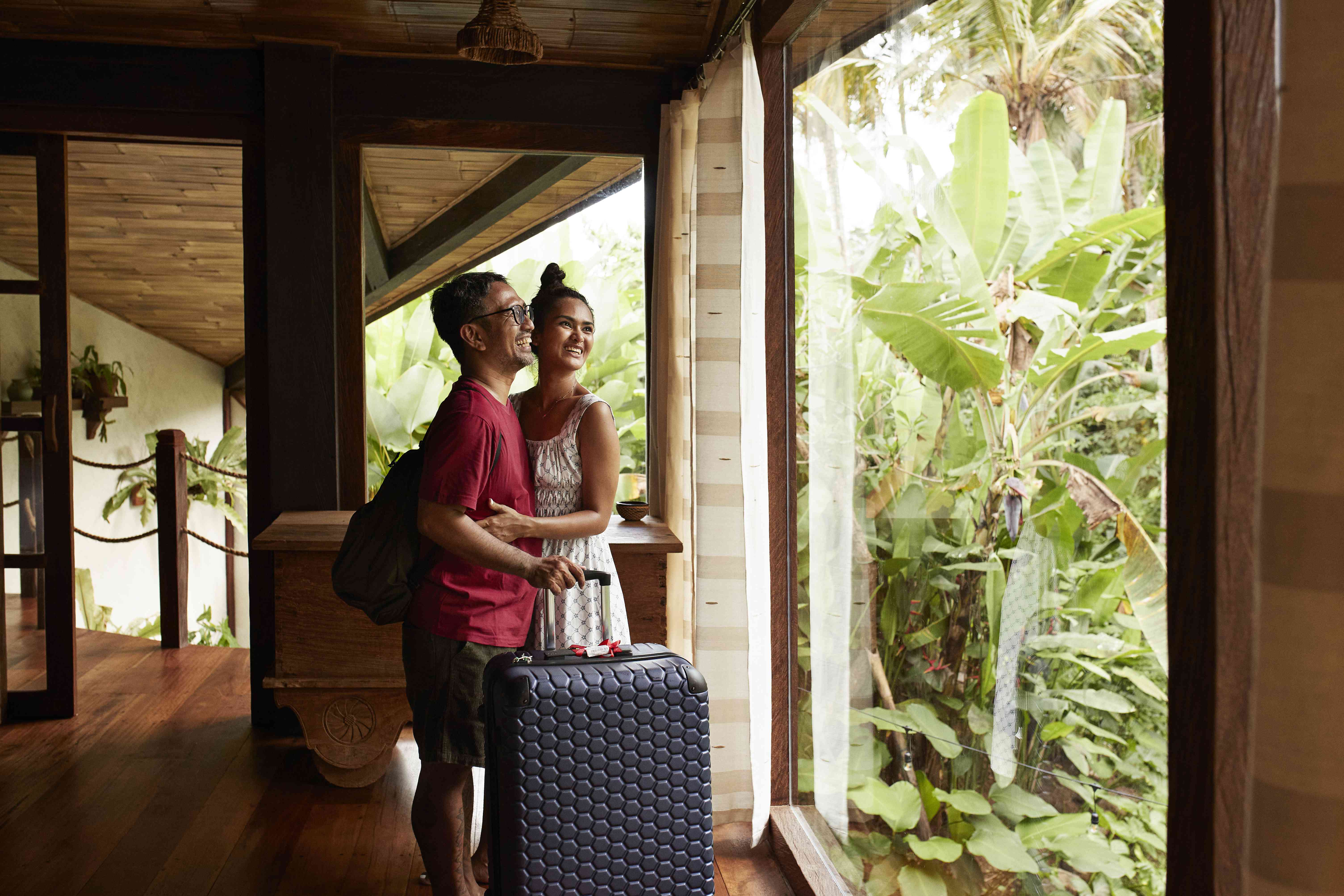 Happy man and woman with luggage standing near window during vacation