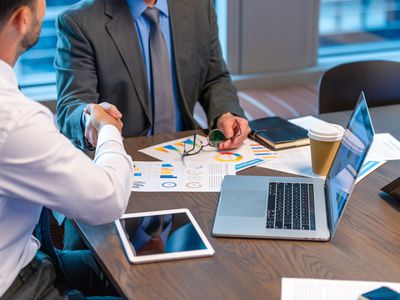 An advisor and client shaking hands while sitting at a table with laptop and tablet on the table.