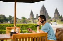 A man sitting outdoors at a restaurant table using a cell phone with the Prambanan temple in the background.