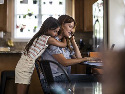 Daughter hugging mom from behind while the other one is on the phone looking for ways to save their house from foreclosure.