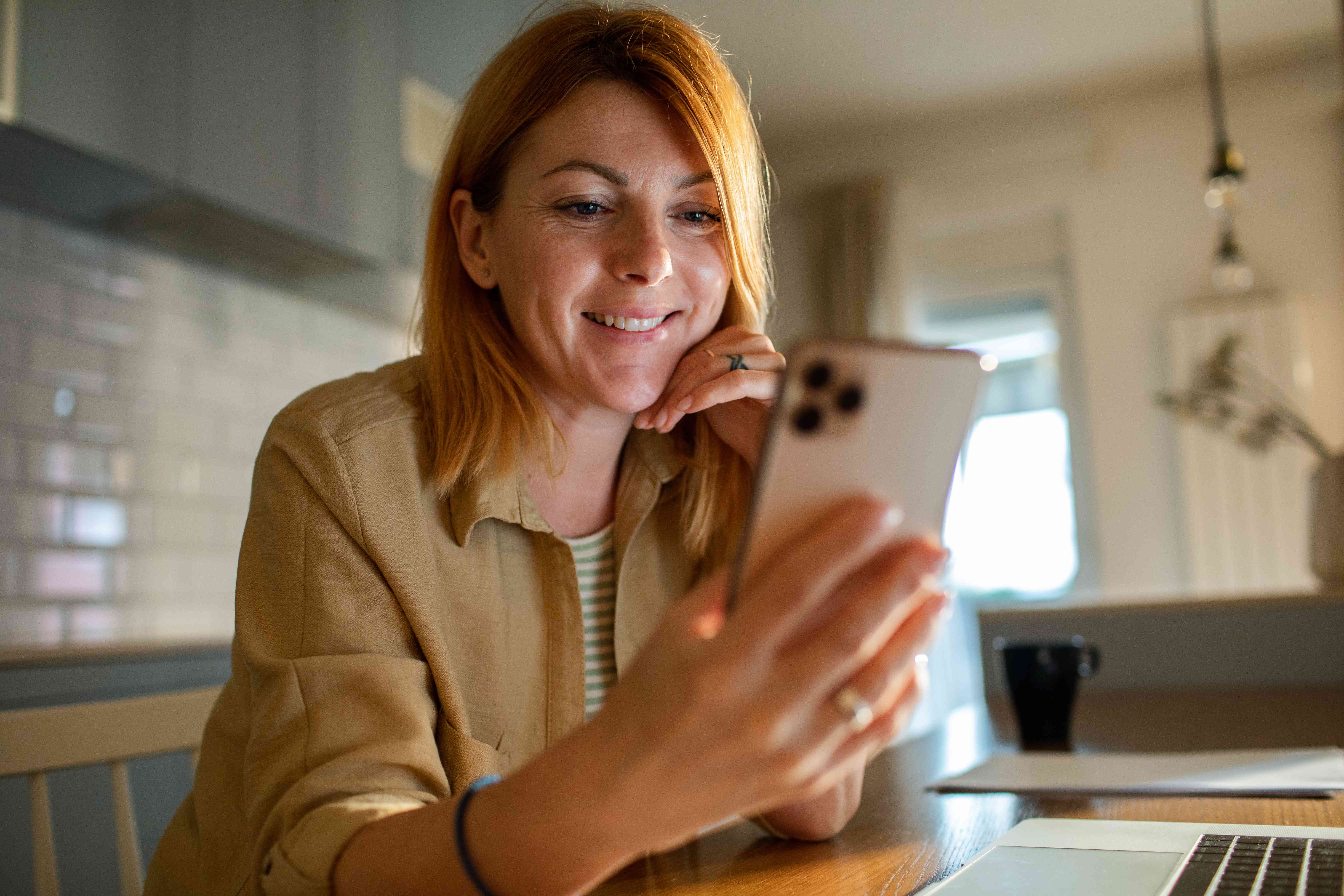 Woman in her 40s sitting in front of a laptop at her kitchen table and smiling as she looks at her smartphone