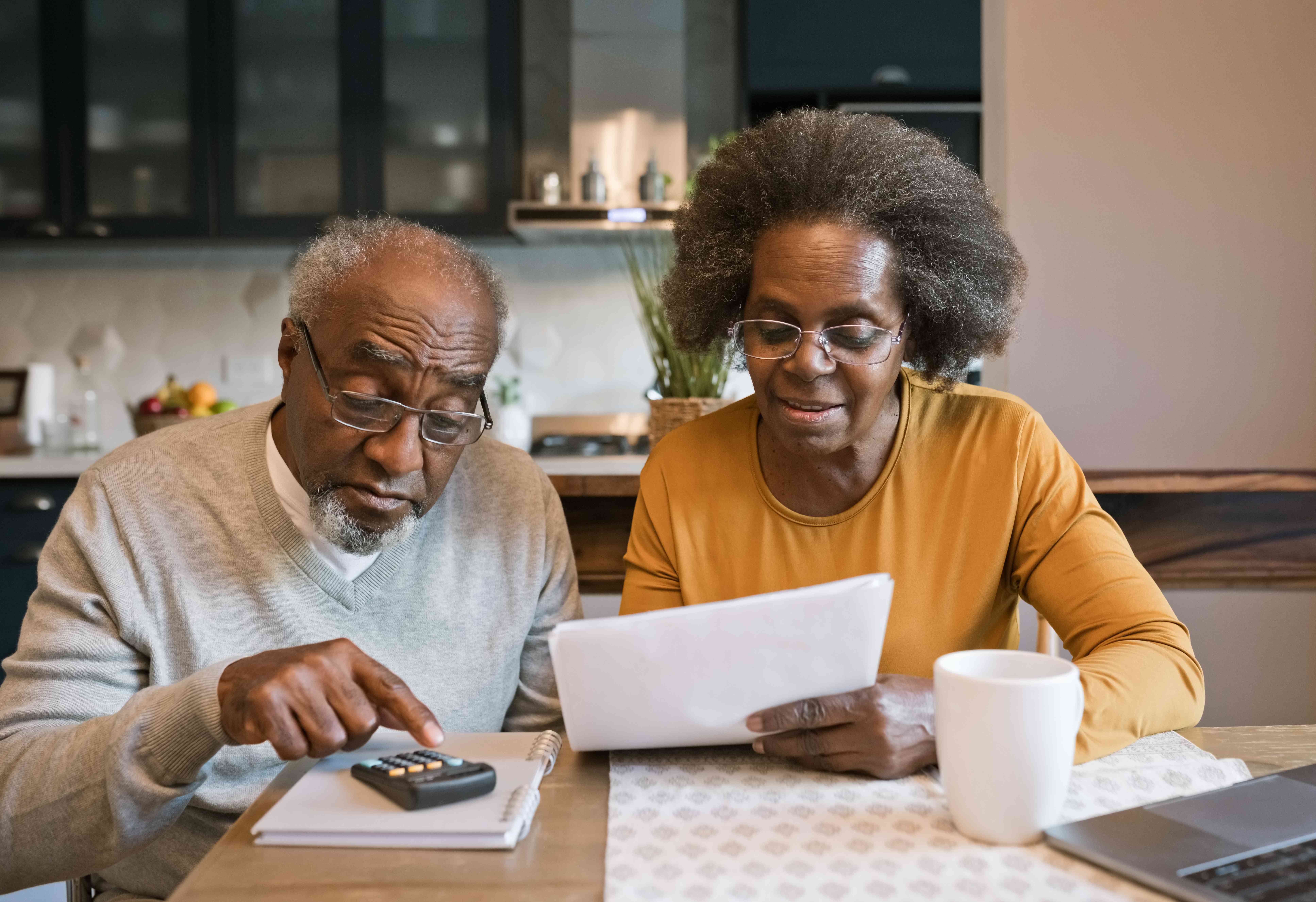Older man and woman sitting together at kitchen table with financial documents, calculator, and laptop