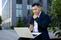 Man sitting on a bench outside an office building, looking at a laptop.