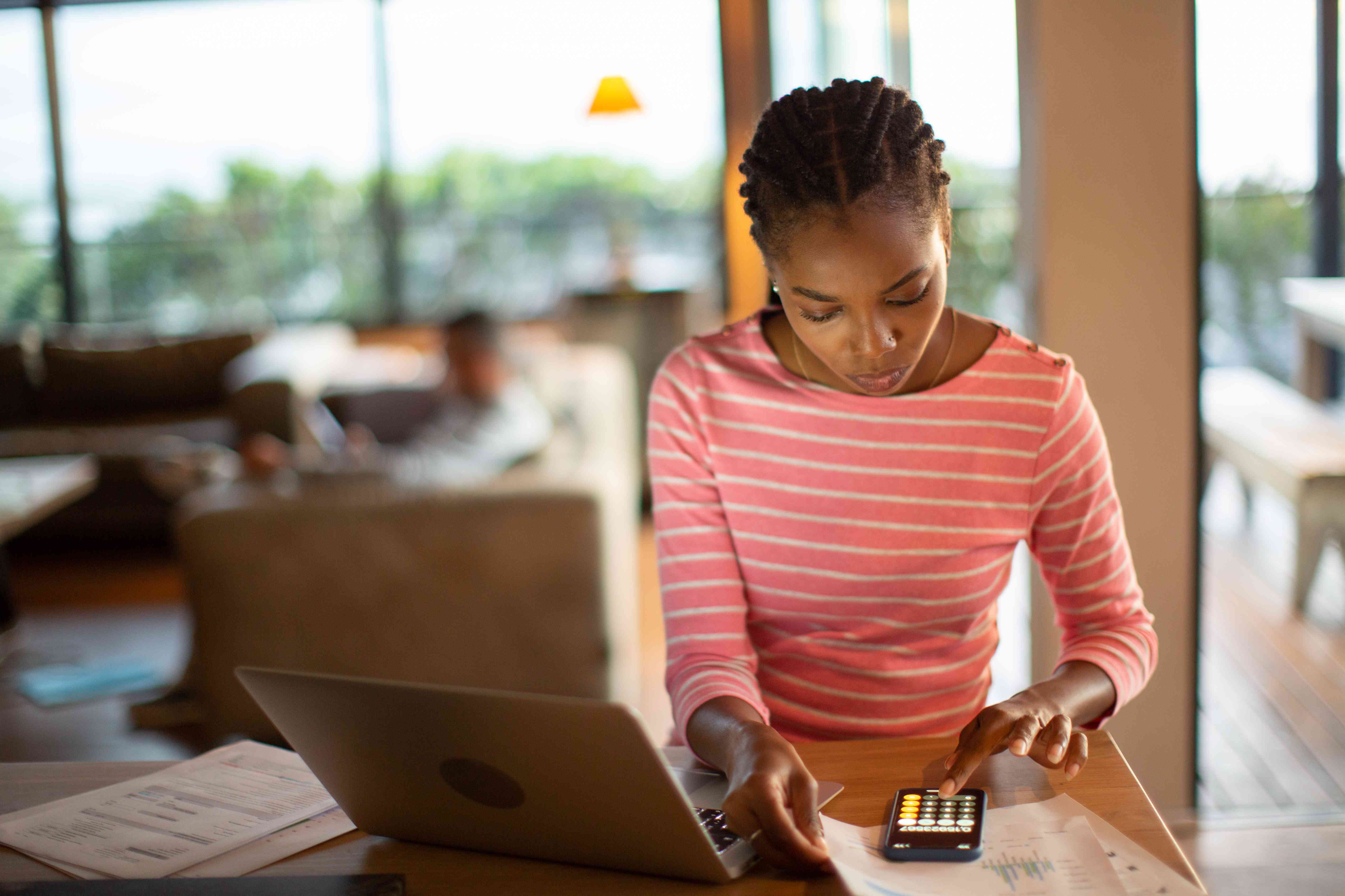 A woman sits at a table with a laptop and a calculator looking at her financial documents.