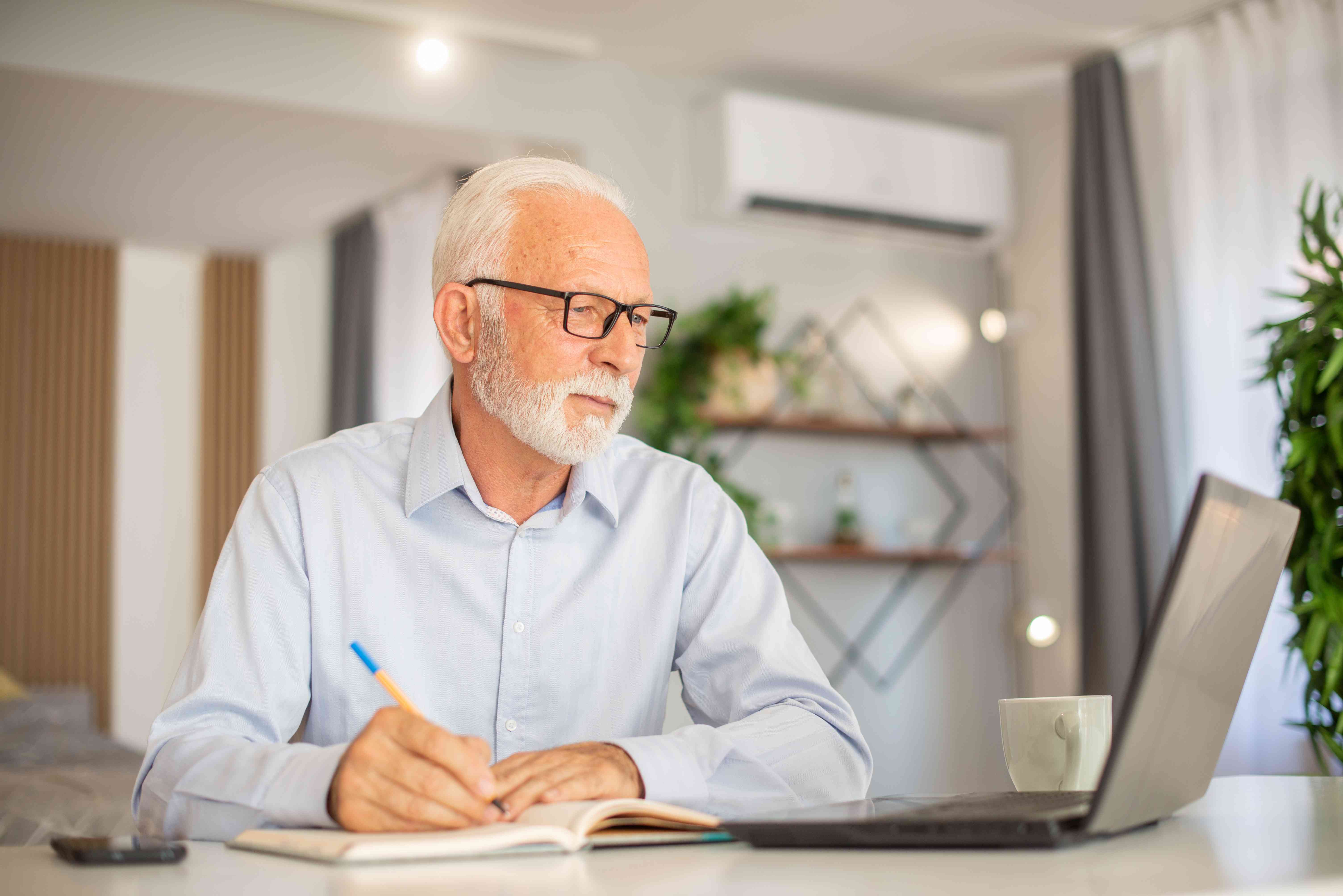 Older man sitting at his kitchen table looking at a laptop and jotting notes in a notebook