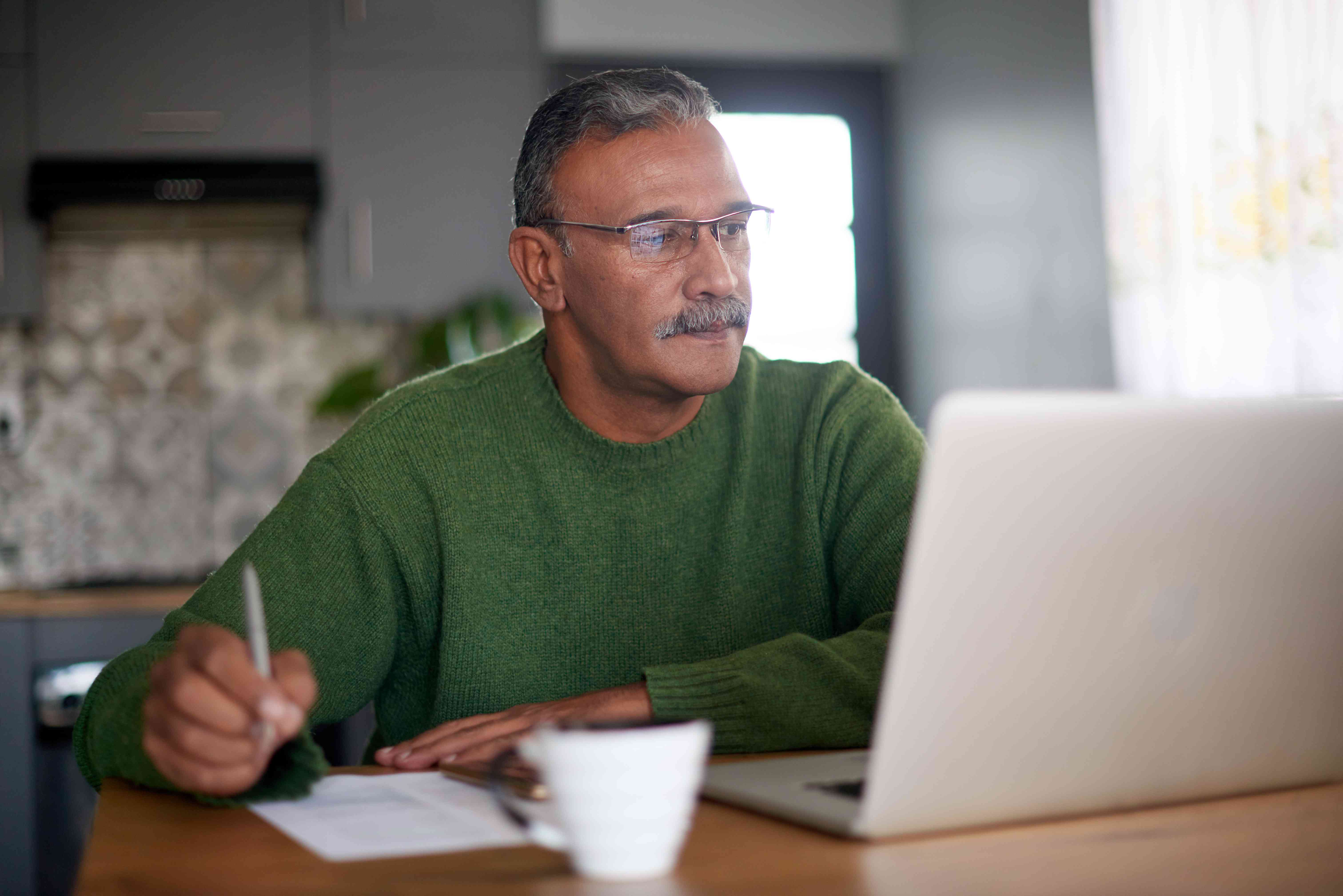 Man in his 50s at his dining room table, looking at a laptop and taking notes