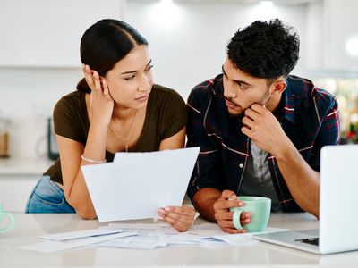 A stressed young couple look at one another, holding papers while drinking coffee in their kitchen, while deciding whether to file a home insurance claim.