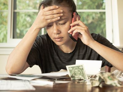 A stressed woman looks at bills on a table, holding a cell phone to her ear.