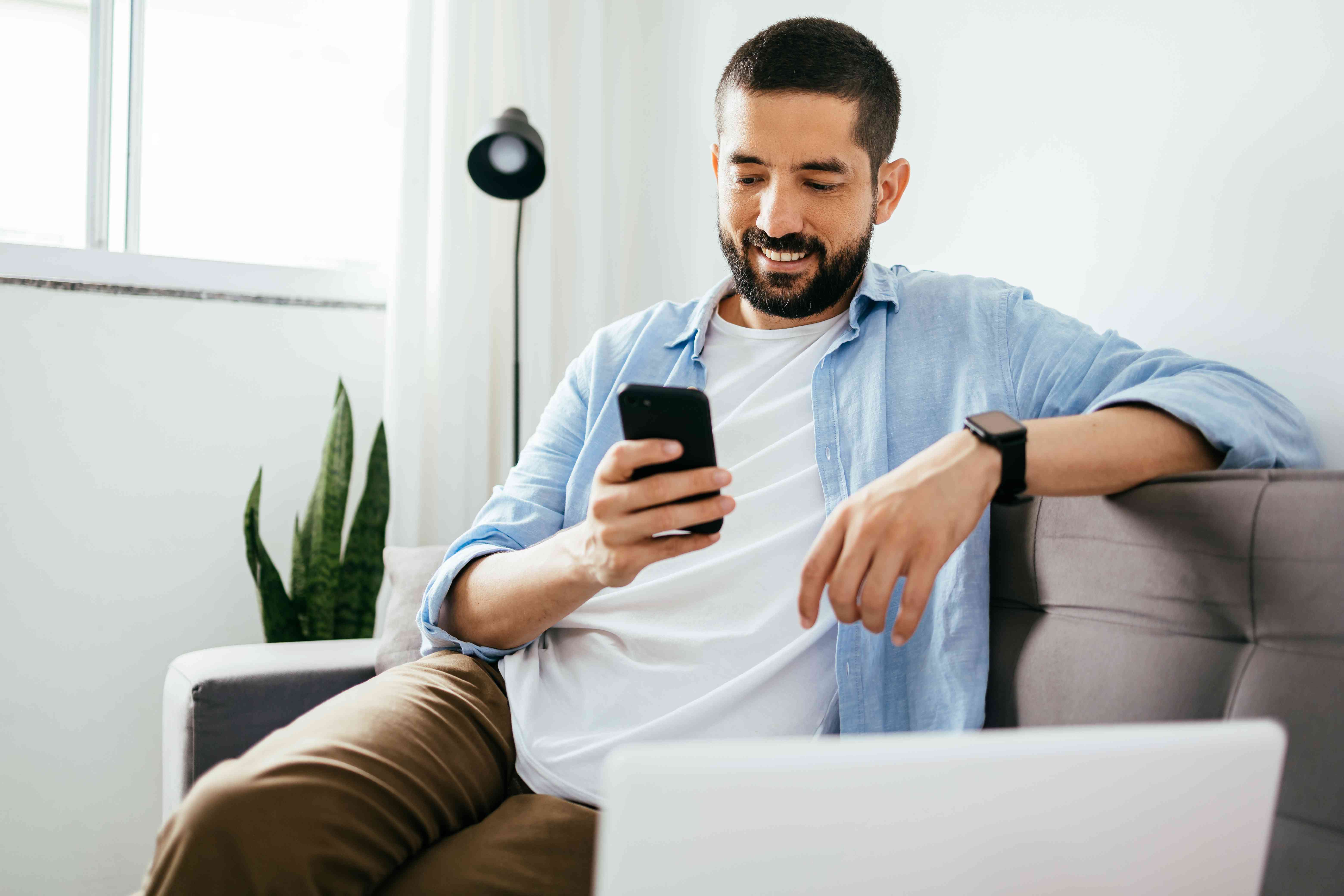 Man in his 30s sitting on his living room couch and looking with a smile at his smartphone