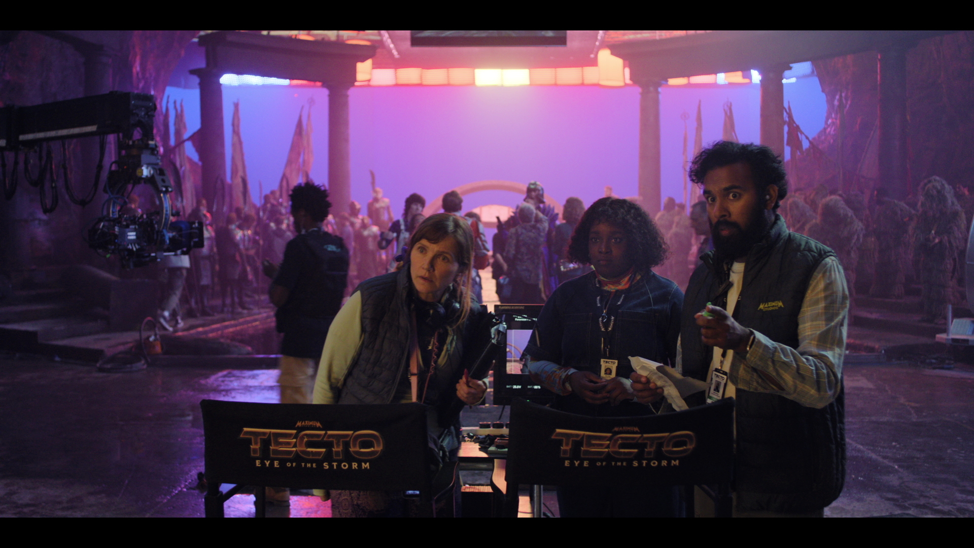 From left to right: Jessica Hynes, Lolly Adefope and Himesh Patel standing behind directors' chairs looking towards camera with a large blue screen set in the background on 'The Franchise'