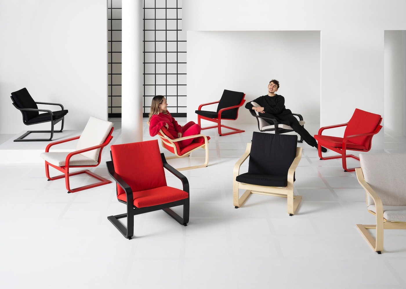 A white room set filled with POÄNG armchairs in different colours. Models dressed in red and black sit in two of the chairs.