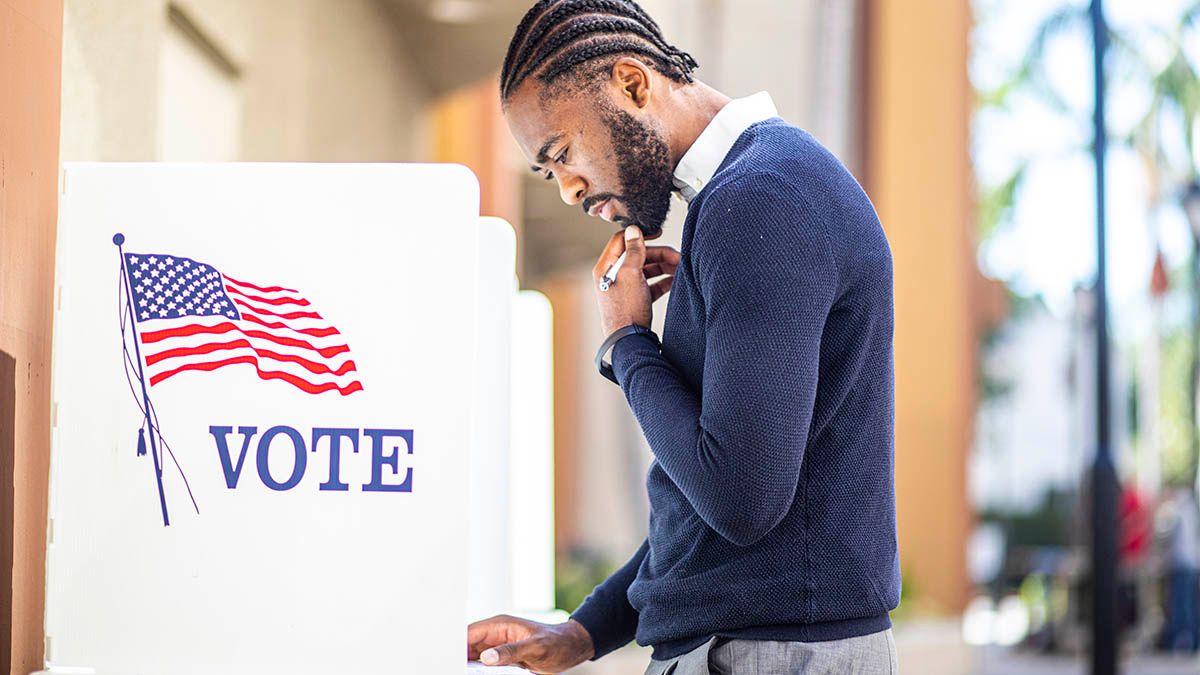 A millenial male ponders his ballot at a ballot box that is decorated with an American flag and the verbiage "vote"