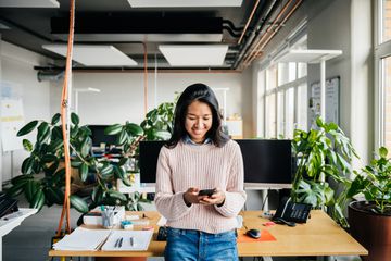 A woman leaning on a desk in an office looking down at her smartphone and smiling