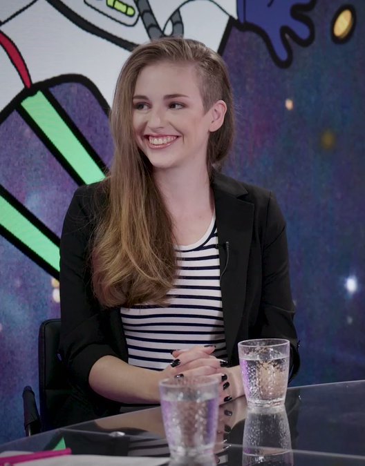 A photograph of Molly White sitting at a table in a video studio. She's      a white woman with long brown hair that's shaved on one side, she has brown eyes, and she's wearing a black and      white striped shirt and black blazer. There is a colorful cartoon background behind her. She's smiling and looking      at someone out of frame.